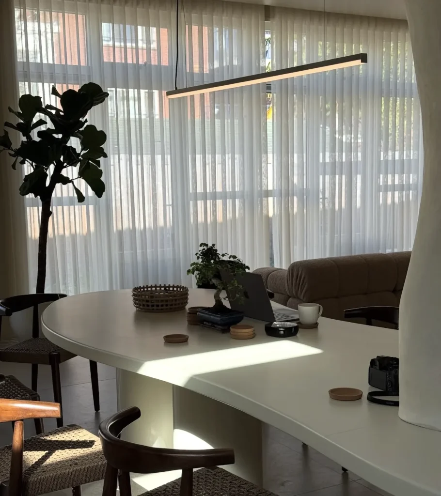 The organic work table by tall curtained windows, with chairs and plants around it