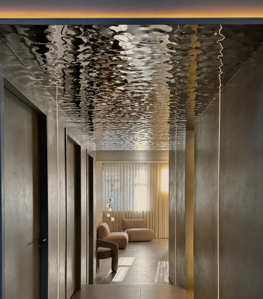 A corridor under a rippled polished steel ceiling leading to a lounge with armchairs