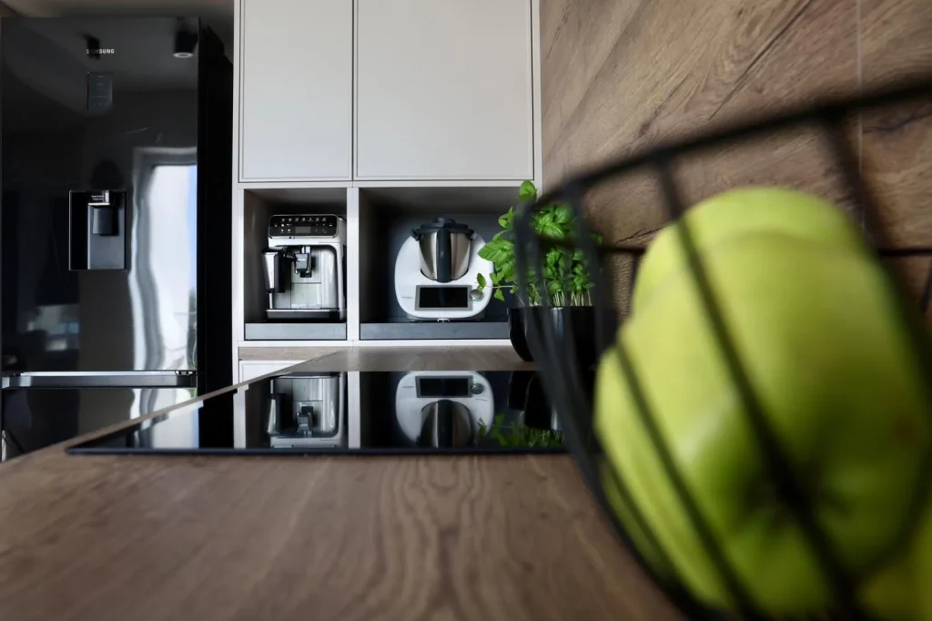 Oak worktop and induction hob leading to a white cabinet niche with a coffee machine and basil, a bowl of green apples in the foreground